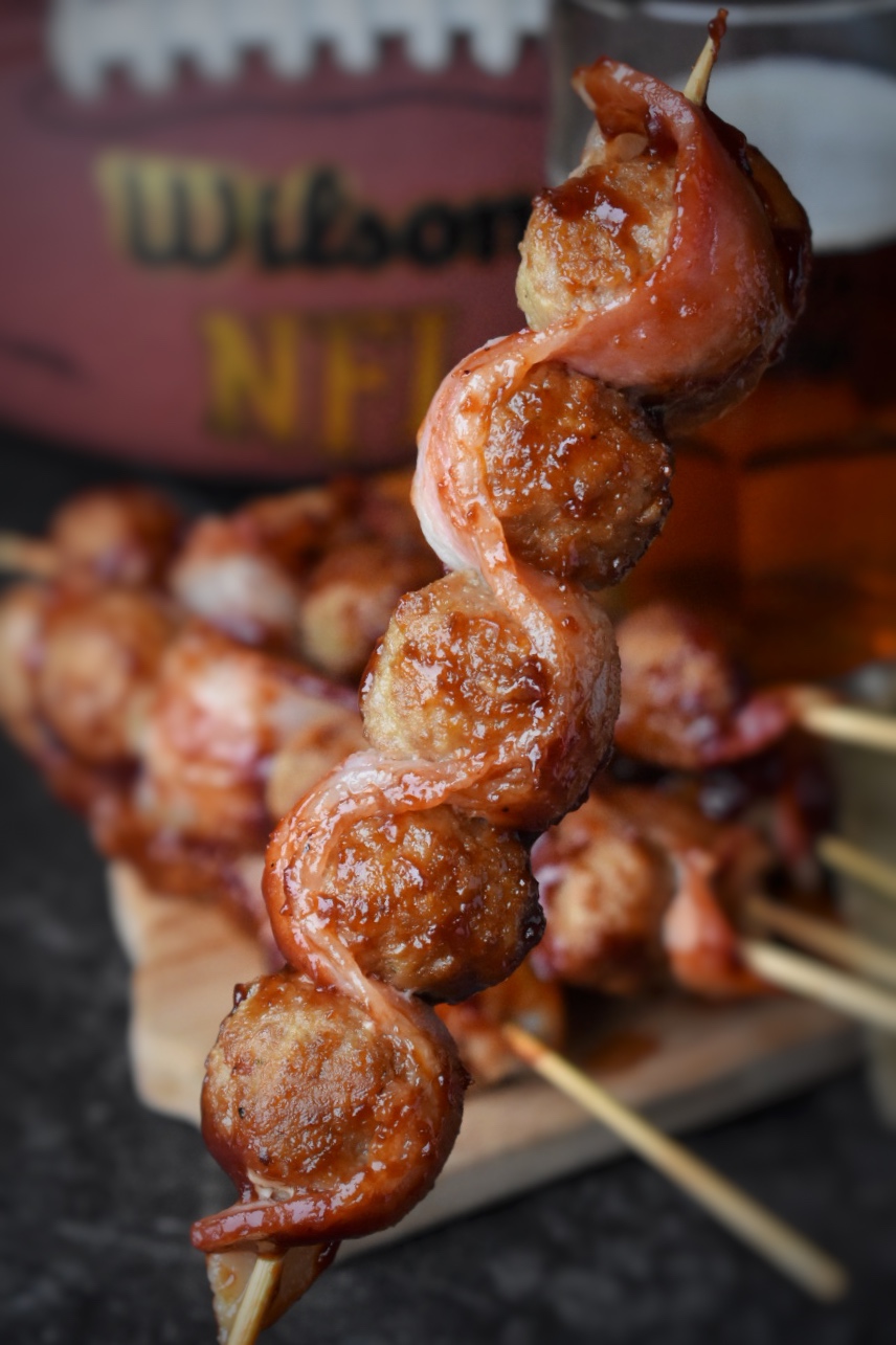 Close up shot of a wooden skewer with meat balls and bacon in front of more skewers in a pile, a football, and a mug of beer.