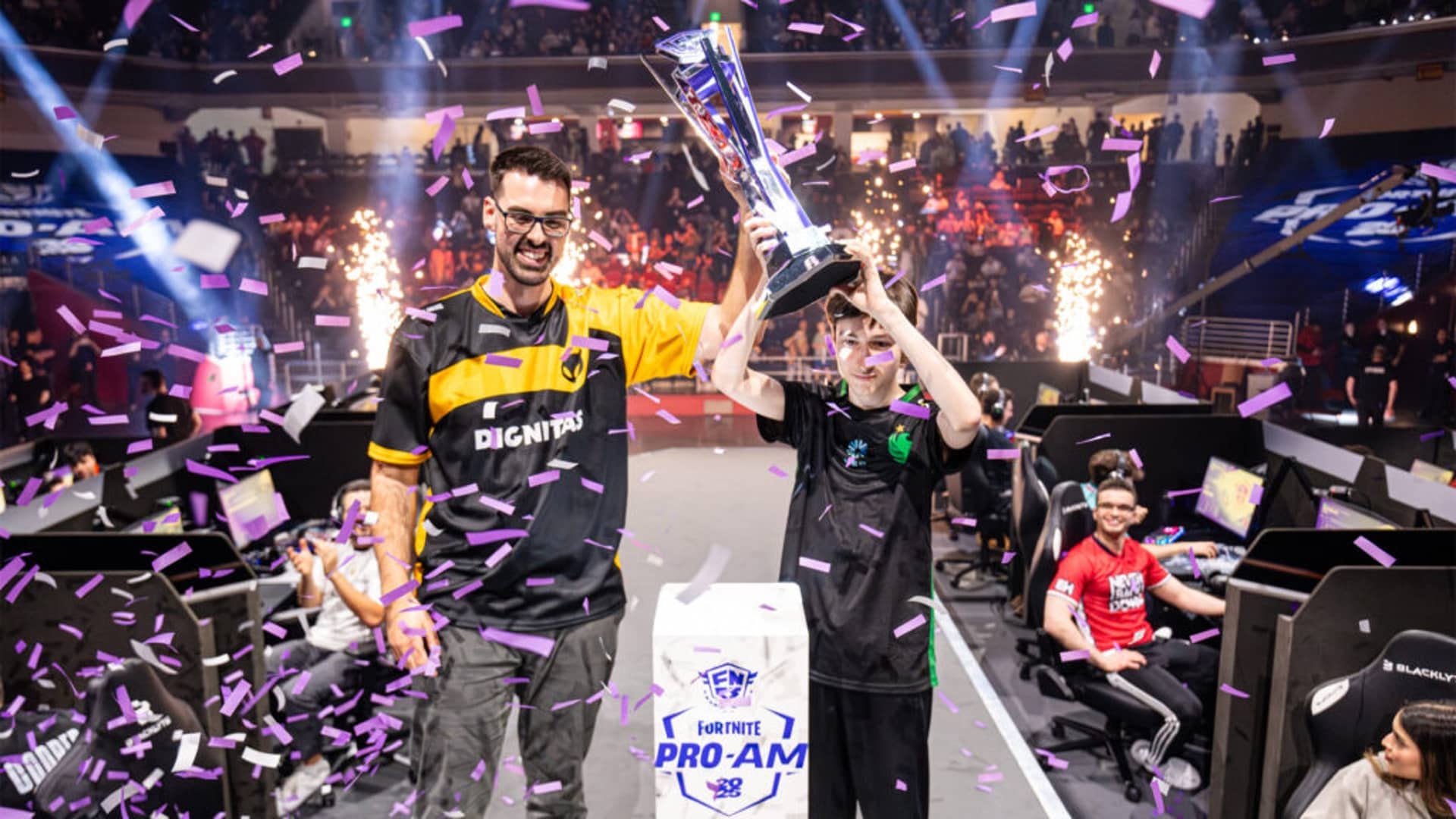 Two people celebrate on stage amid purple confetti at a gaming event. One holds a trophy high, both smiling. A cheering crowd and fireworks are visible