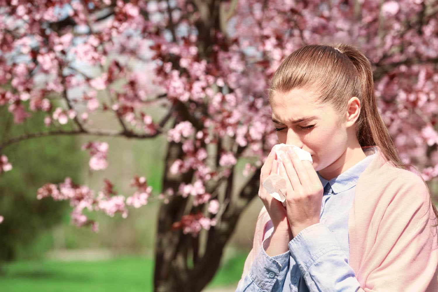 Sneezing young girl with nose wiper among blooming trees in park