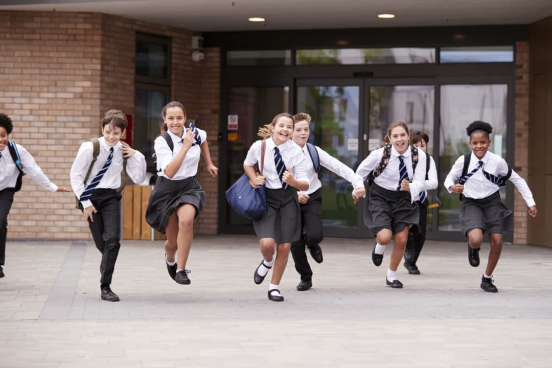 Group Of High School Students Wearing Uniform Running Out Of School Buildings Towards Camera At