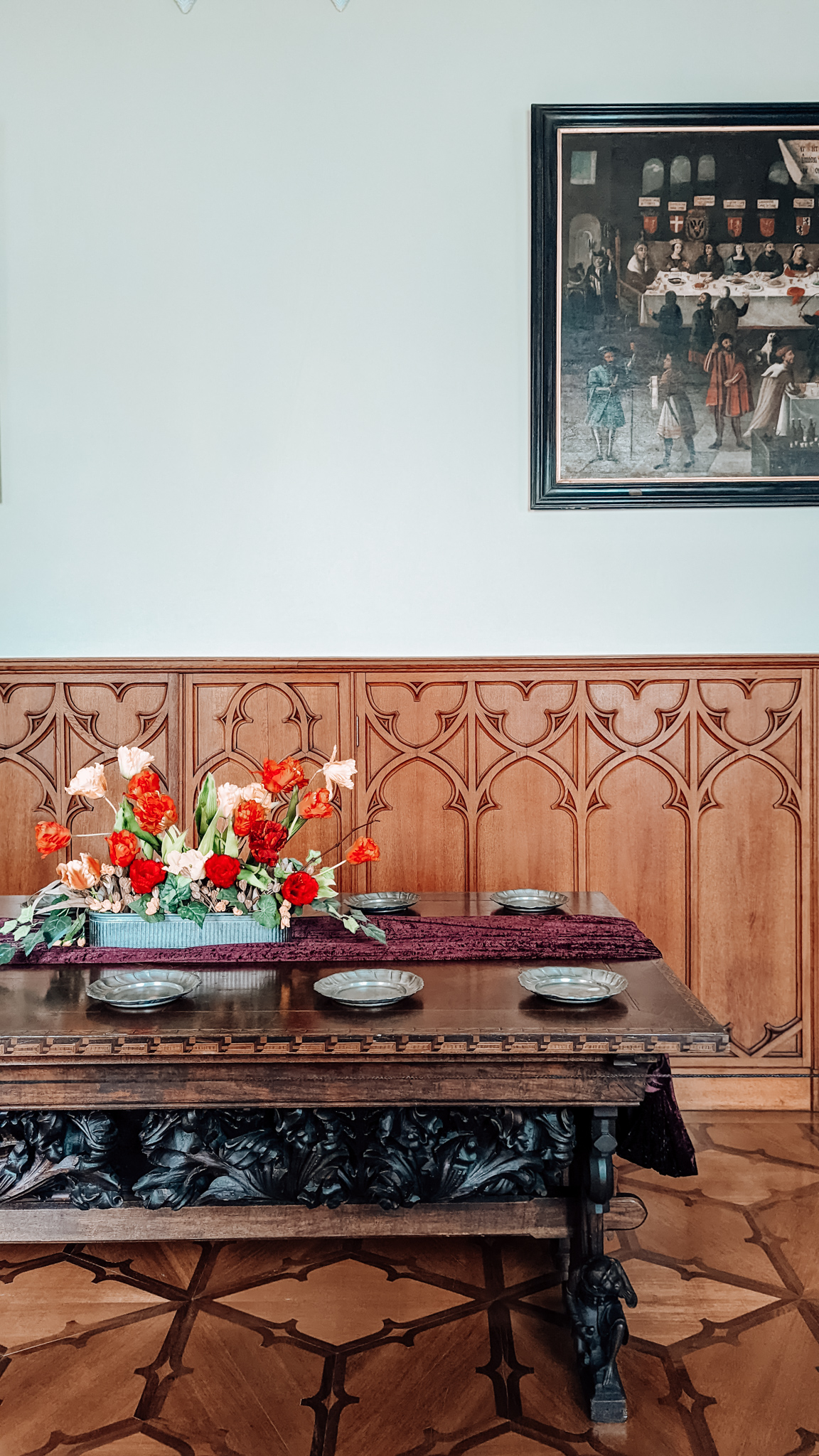 The right end of a long table which is set up to represent how it looked when used for dining. In the centre is a large bouquet of flowers. Above it and to the right is part of a painting hanging on the wall.