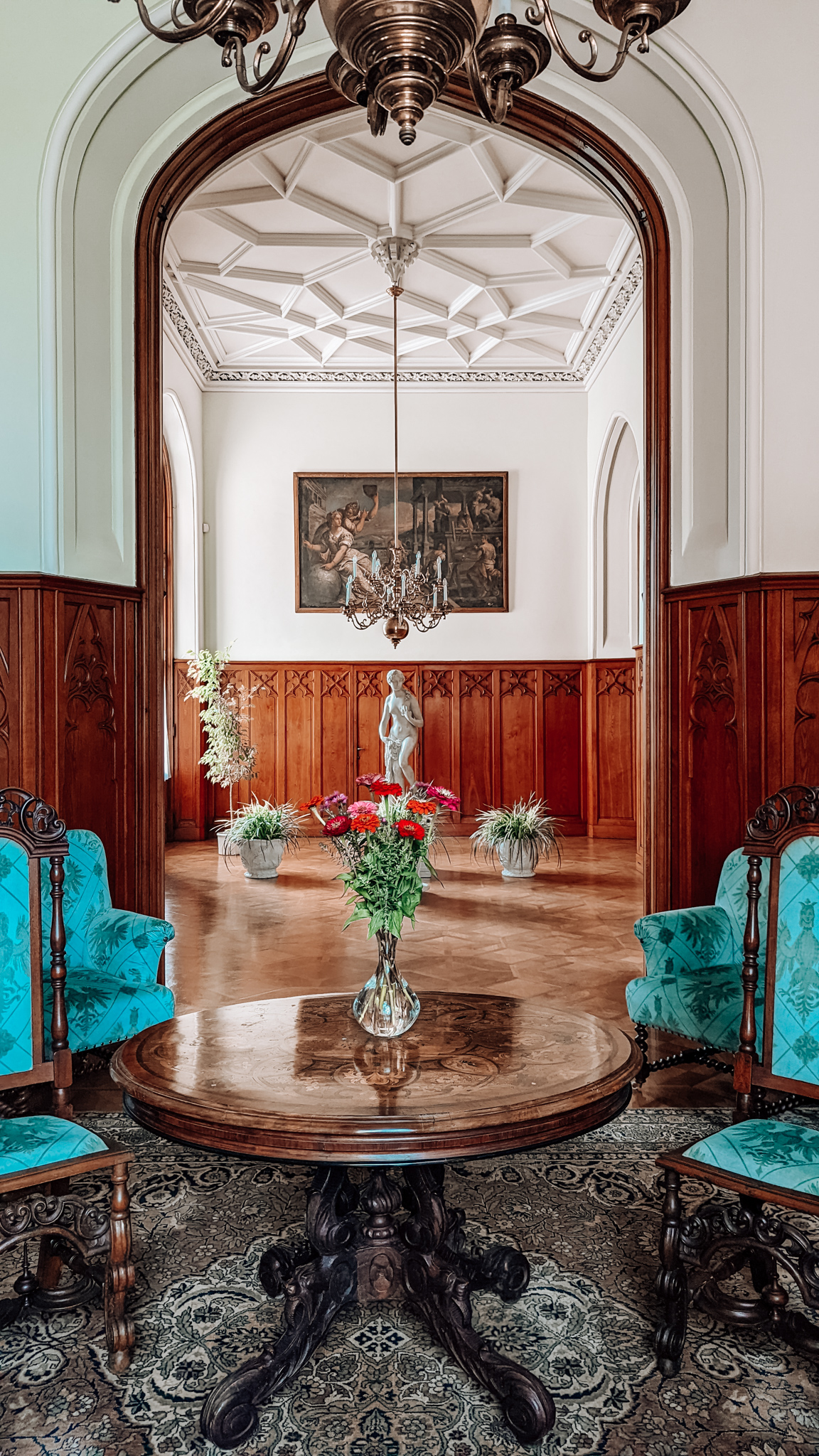 A small table sits in the forefront of the photo, with red flowers in a vase. Either side of the table are two bright blue vintage chairs. Behind this, there is a stone statue of a lady in the background of another room, with a painting hanging on the wall.