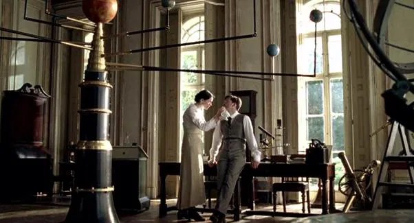 A scene from Einstein and Eddington where Rebecca Hall and David Tennan in period clothing stand close together near a window, surrounded by scientific equipment.
