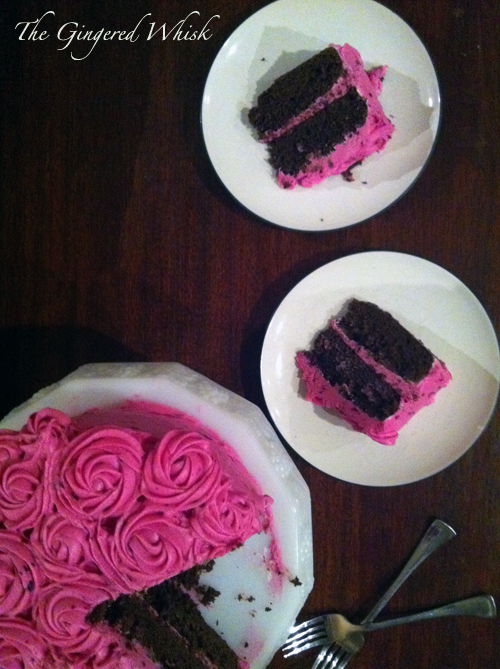 A vertical top down image of a chocolate cake with pink frosting and two slices set on white plates on a wooden surface.