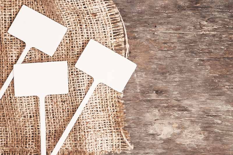 Three white plastic garden signs on a rustic woven fabric, on a wooden surface.