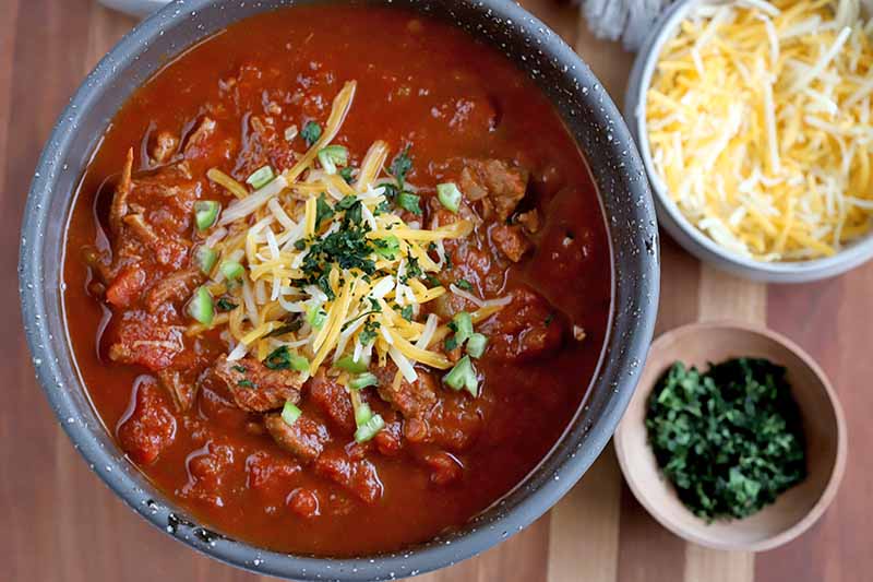 A close up top down picture of a bowl of chili, sprinkled with grated cheese and herbs, set on a wooden surface, with small bowls of cheese and herbs to the right of the frame in soft focus.