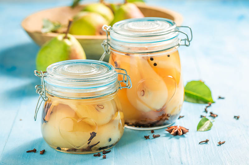A close up horizontal image of two jars filled with canned fresh pears set on a blue wooden surface with ripe fruits in a wooden bowl in the background.