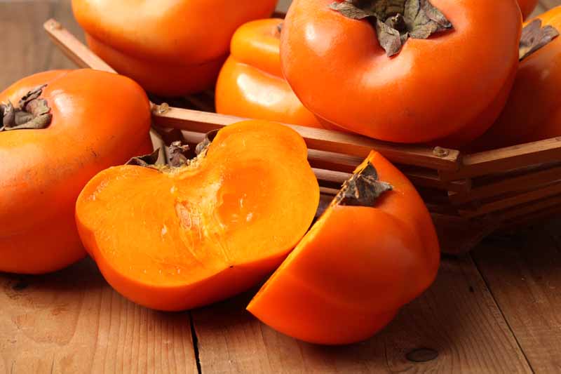 A close up horizontal image of Diospyros kaki fruit in a wooden bowl with one sliced in half to show the orange flesh, set on a wooden surface.