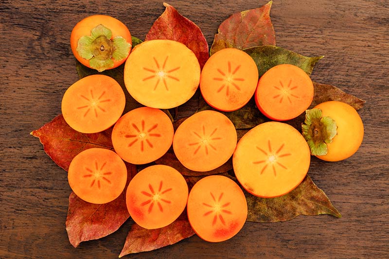 A close up horizontal image of freshly harvested persimmons sliced in half to show the orange flesh inside, set on a wooden surface.