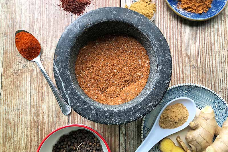 A close up top down horizontal image of a mortar filled with ras-el-hanout spice blend set on a wooden surface with individual spices surrounding.
