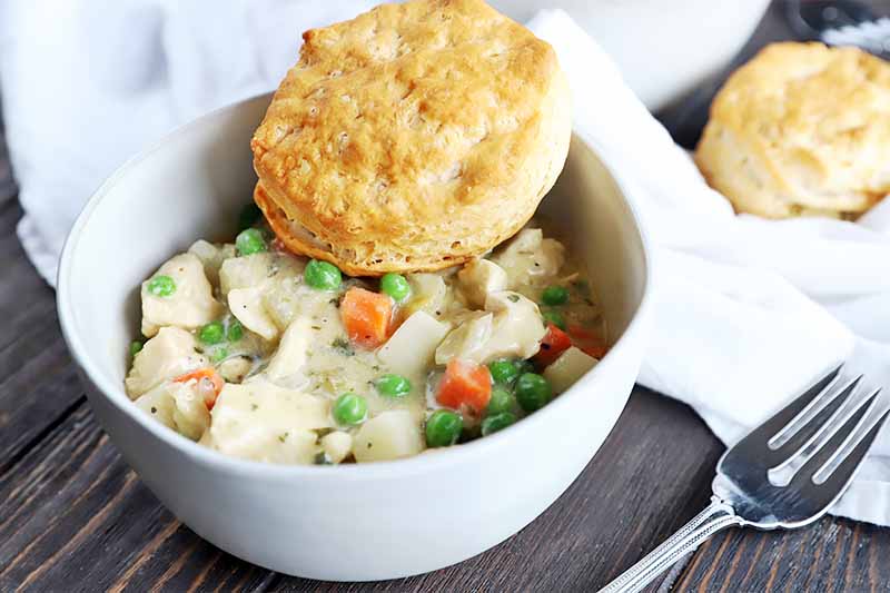 A close up horizontal image of a white bowl with freshly prepared chicken pot pie set on a wooden surface with a fork and white cloth to the left of the frame.