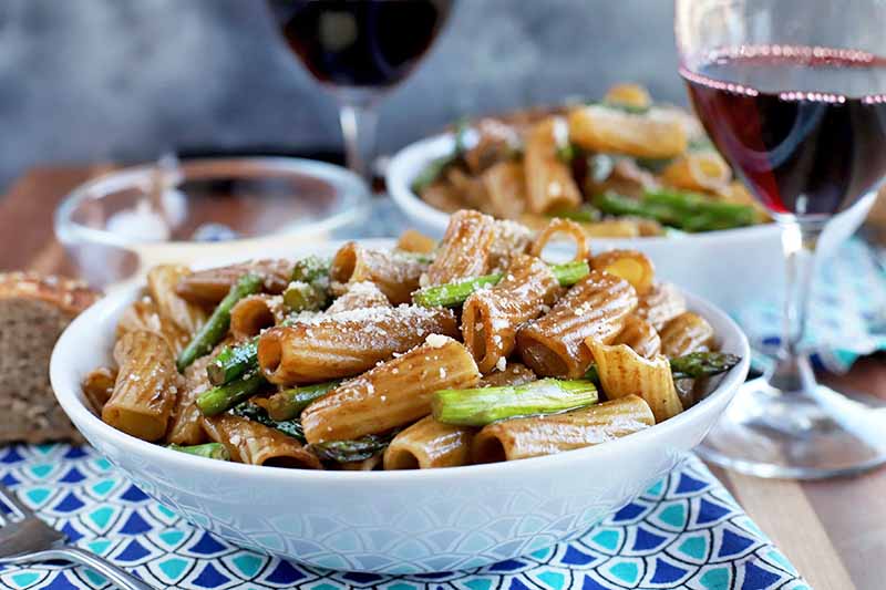 A close up horizontal image of a white bowl filled with a fresh pasta dish with a wine glass in the background.