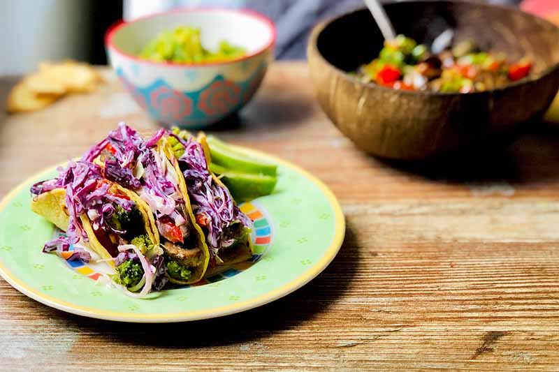 A close up horizontal image of a plate of garlic and ginger tacos served with slaw set on a wooden surface, with a wooden bowl in the background in soft focus.