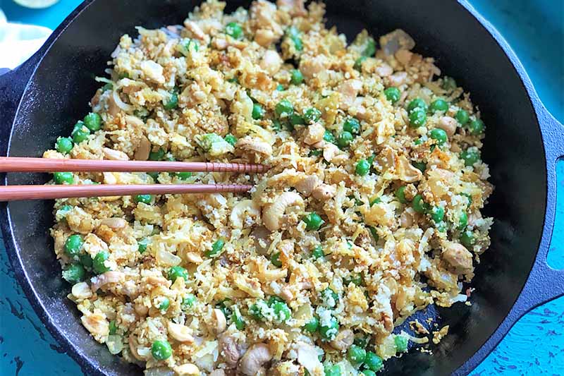 A close up horizontal image of cauliflower fried rice with vegetables in a black metal bowl with chopsticks to the left of the frame.