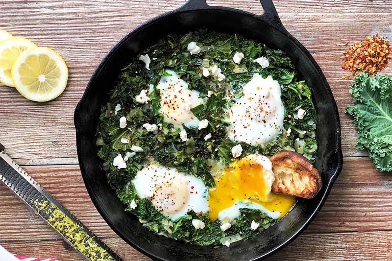 A close up horizontal image of a skillet with freshly prepared kale and eggs set on a wooden surface.