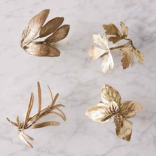 A close up square image of four napkin rings in the shape of herbs set on a marble surface.