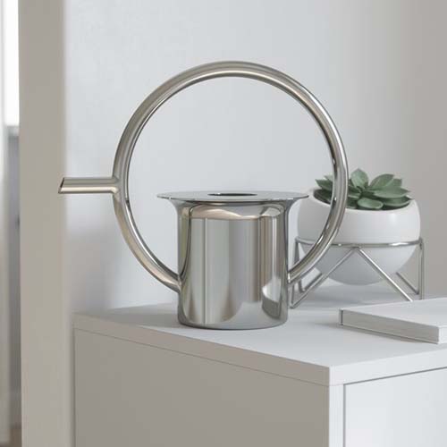 A close up square image of a modern Umbra Quench Stainless Steel watering can set on a white surface with a succulent houseplant in the background.