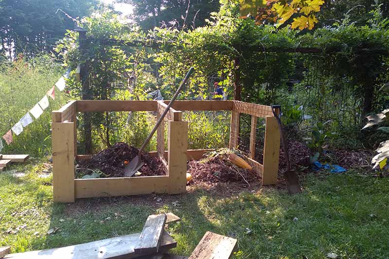 A close up horizontal image of two wooden compost piles pictured in light filtered sunshine.