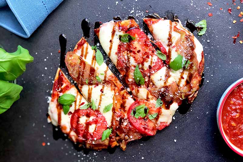 A close up horizontal image of a freshly prepared caprese naan pizza drizzled with balsamic on a dark gray surface.