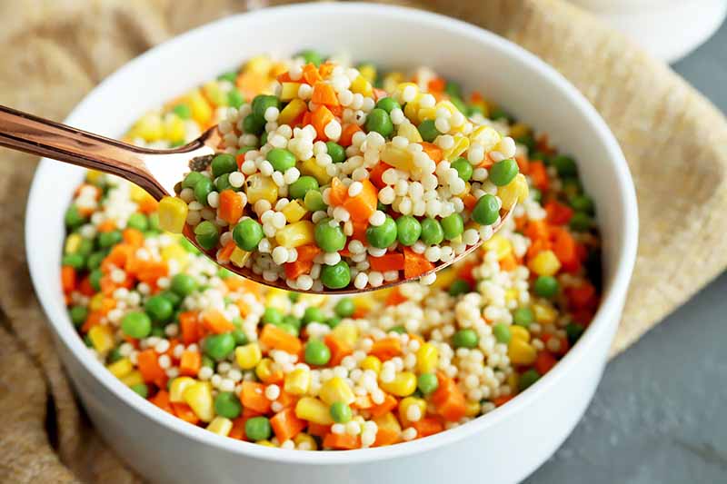 A close up of a bowl of citrus vegetable couscous set on a wooden surface.