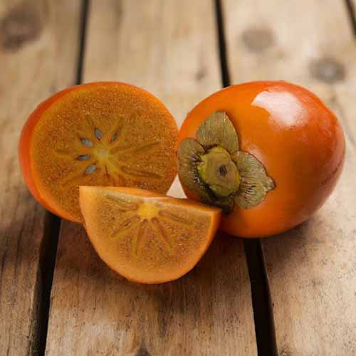 A close up of a whole and sliced &lsquo;Chocolate&rsquo; persimmon set on a wooden surface.