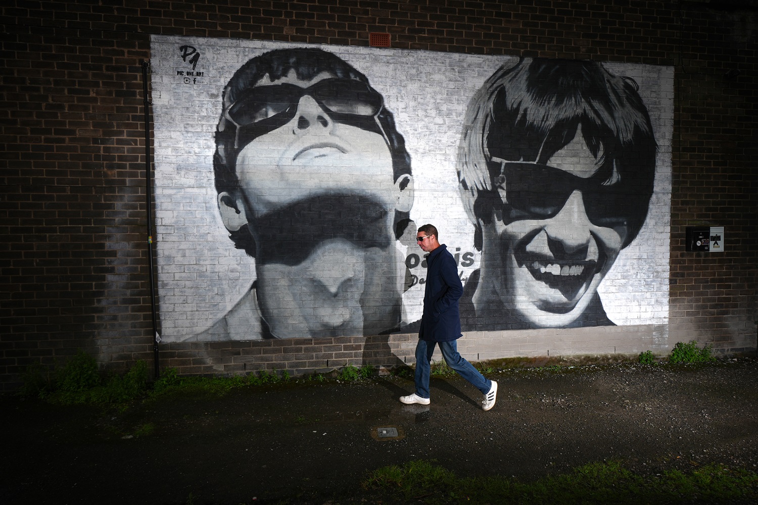An Oasis fan is photographed next to a new mural depicting Liam and Noel Gallagher of Oasis, created by Manchester street artist Pic.One.Art., on the side of the Sifters Record store in Burnage on August 28, 2024 in Manchester, England.