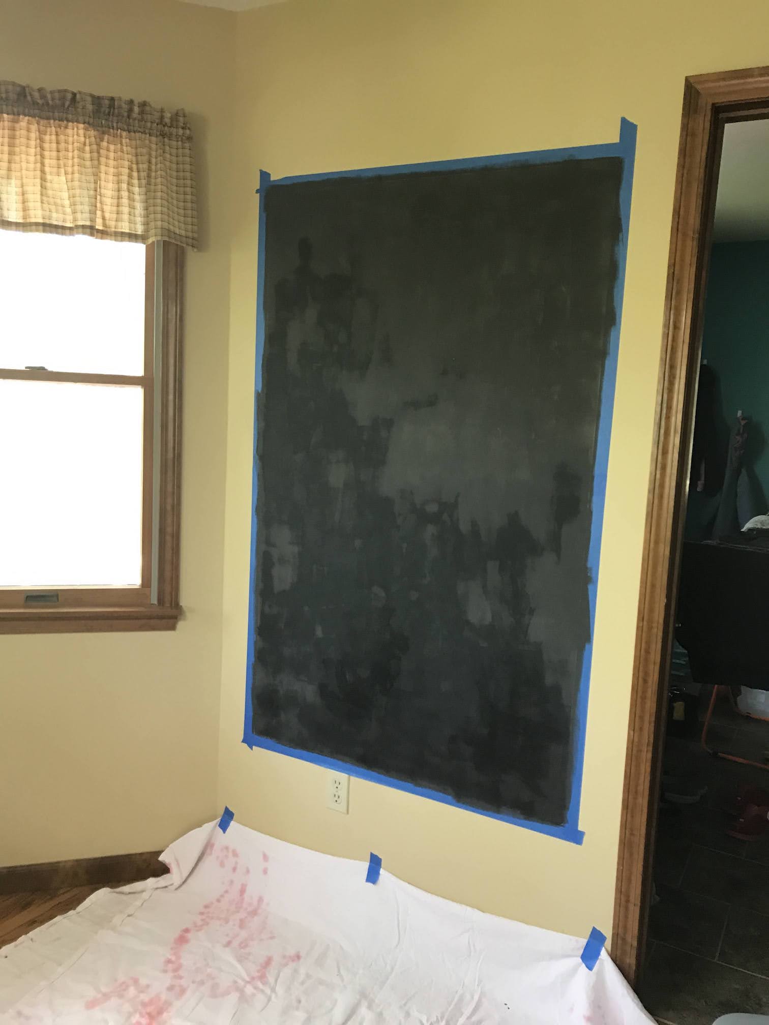 DIY chalkboard wall in progress with black paint on wall outlined with painter's tape next to a window