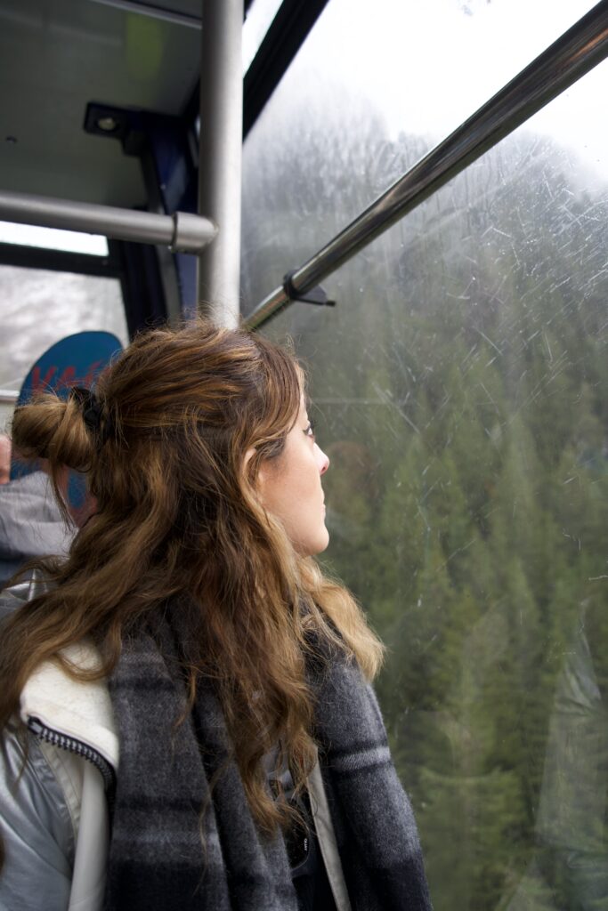 Woman with brown hair wearing a tartan scarf and white jacket sits in a cable car staring into the distance