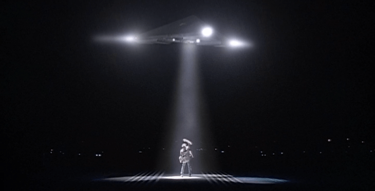 A triangular spacecraft is seen hovering above Mulder, who is looking up at it with an arm raised over his head.