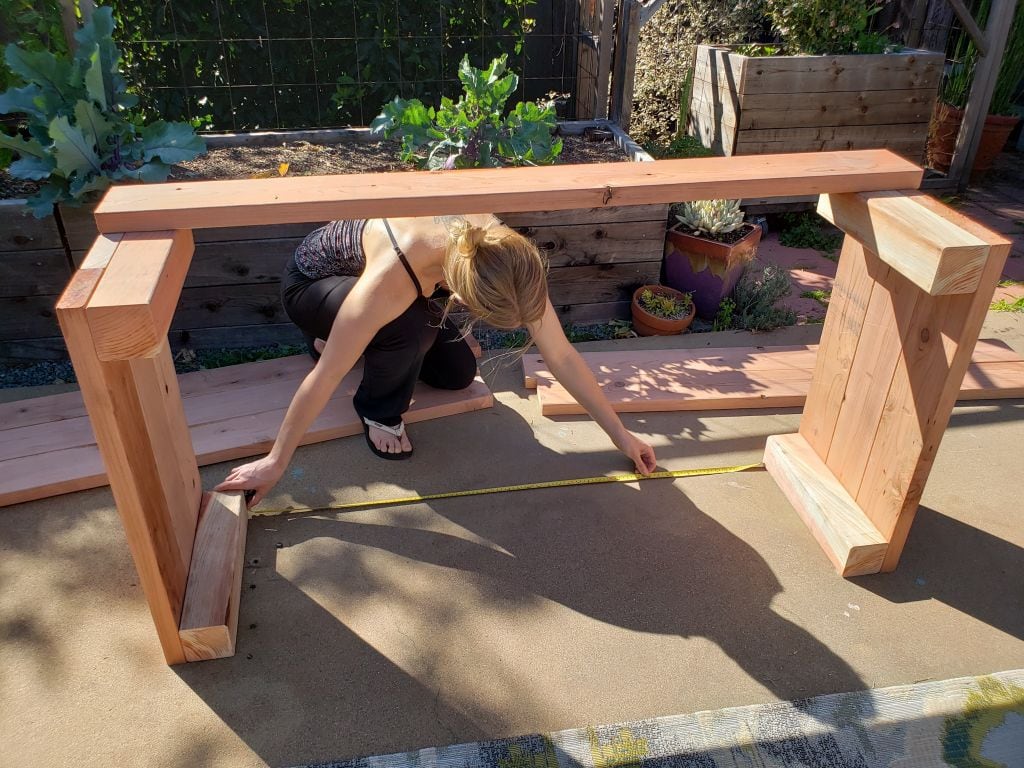 DeannaCat is shown kneeling down taking a measurement of a partially made wooden raised garden bed. Constructing garden beds and structures may be a necessity when on decides to start a homestead.