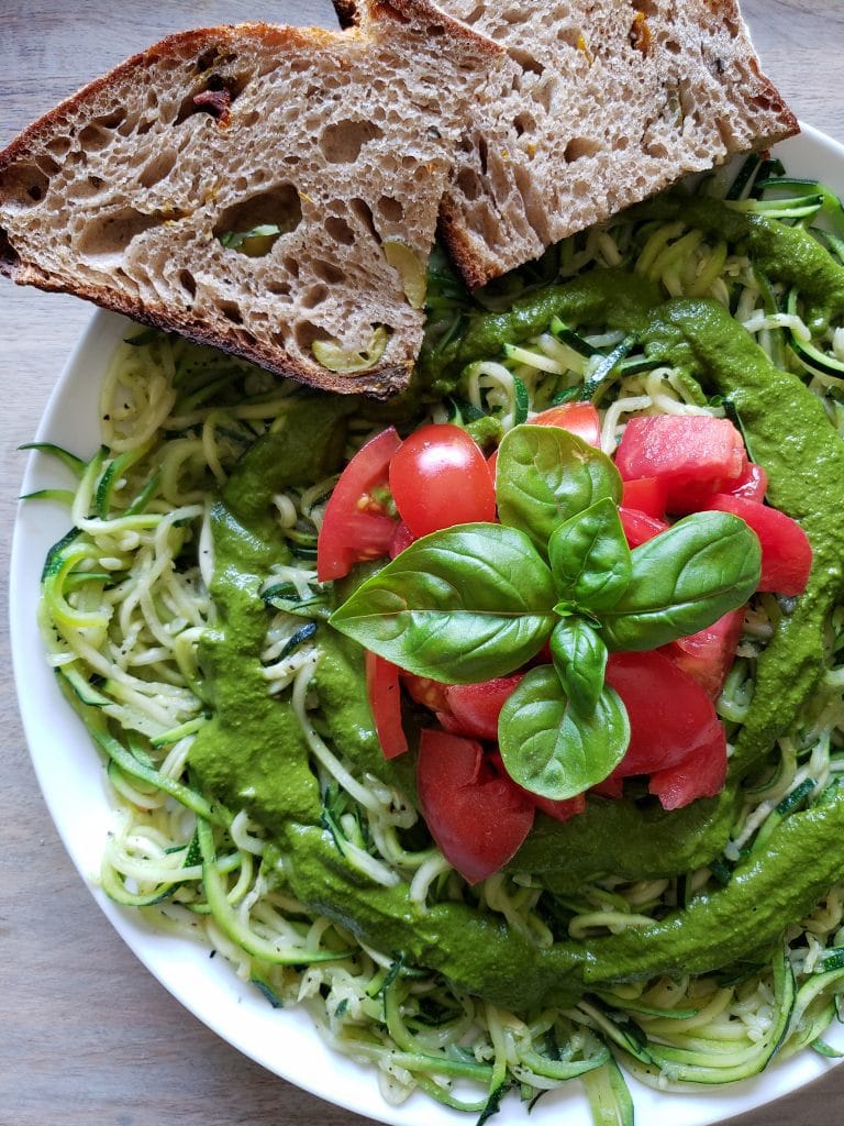 The final plating of zoodles is shown on a plate which contains a bed of zoodels, drizzled with fresh pesto, a nice pile of fresh tomatoes is placed in the middle of the pile along with a sprig of basil. Two slices of fresh sourdough bread are sitting on the edge of the plate.