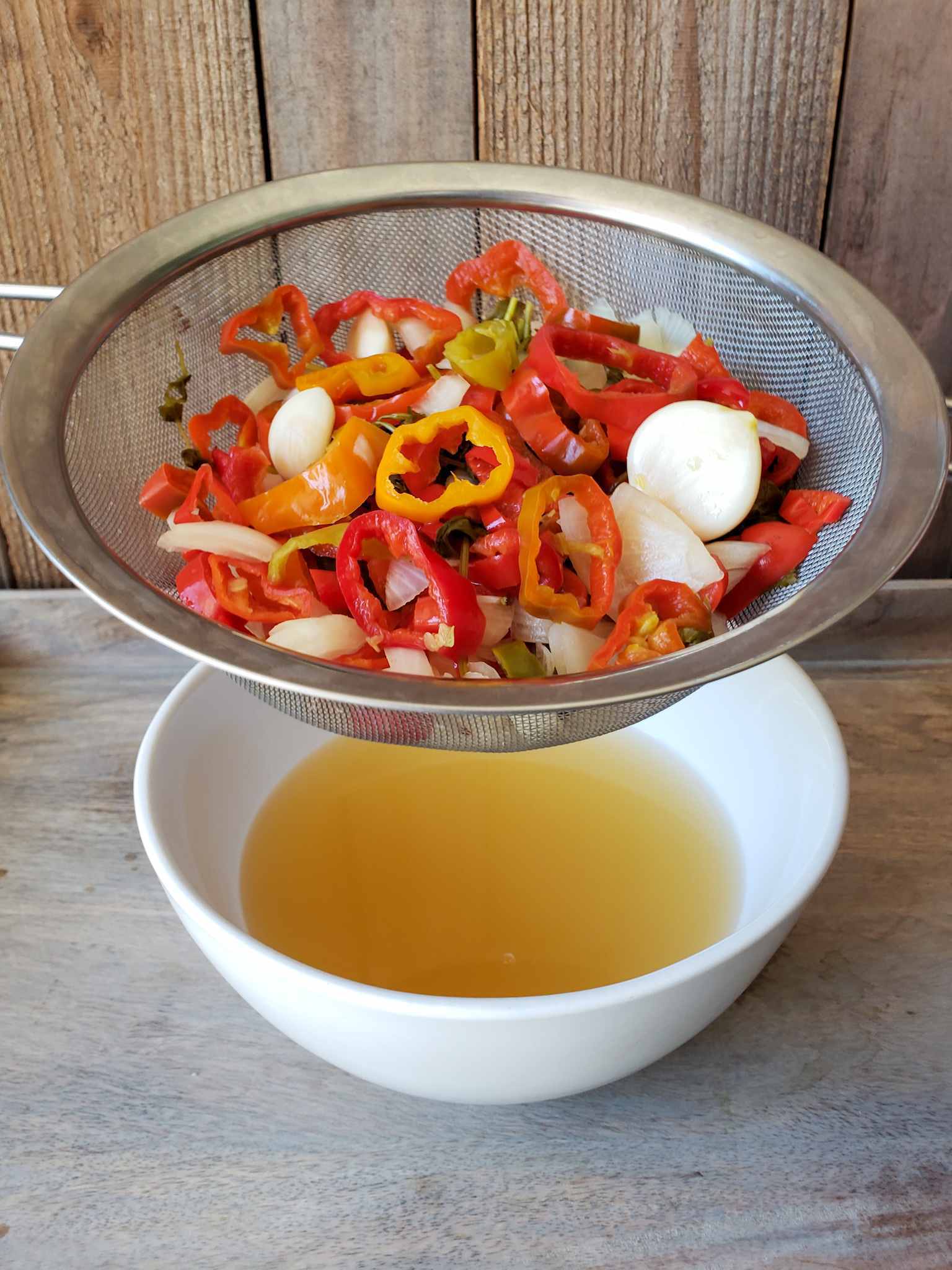A stainless steel strainer full of fermented peppers, garlic, and onions is hovering over a white below below. The bowl holds the drained cloudy yellow brine from the fermentation process, which a portion will be blended back with the peppers to create the final fermented hot sauce.
