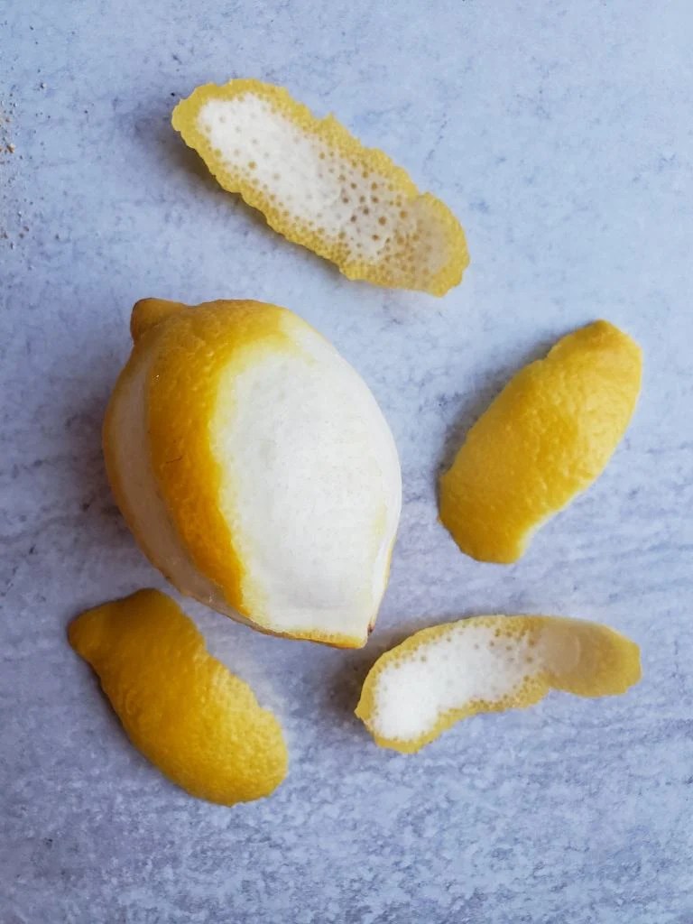 A lemon sits atop a washed concrete surface, it has been partially peeled with a household peeler and the lemon peels are laying around the perimeter of the lemon and its now mostly pithy white exterior. The lemon peels will be soon turned into lemon powder.