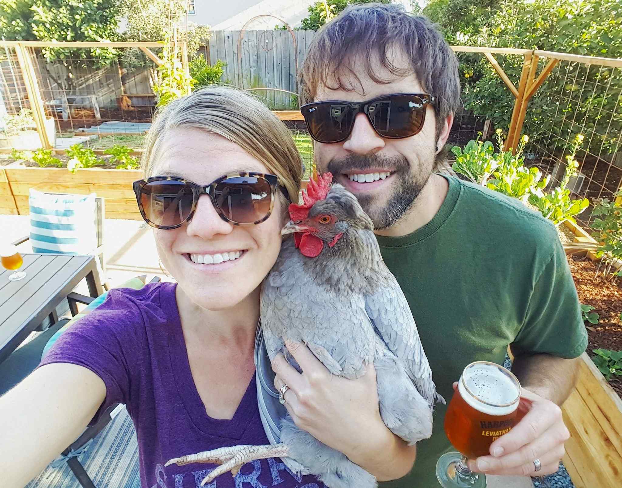 DeannaCat and Aaron standing in their newly designed and constructed patio garden. DeannaCat is holding a grey chicken and Aaron is holding a glass of beer. Vegetable plants have been planted and can be seen in the new garden beds.
