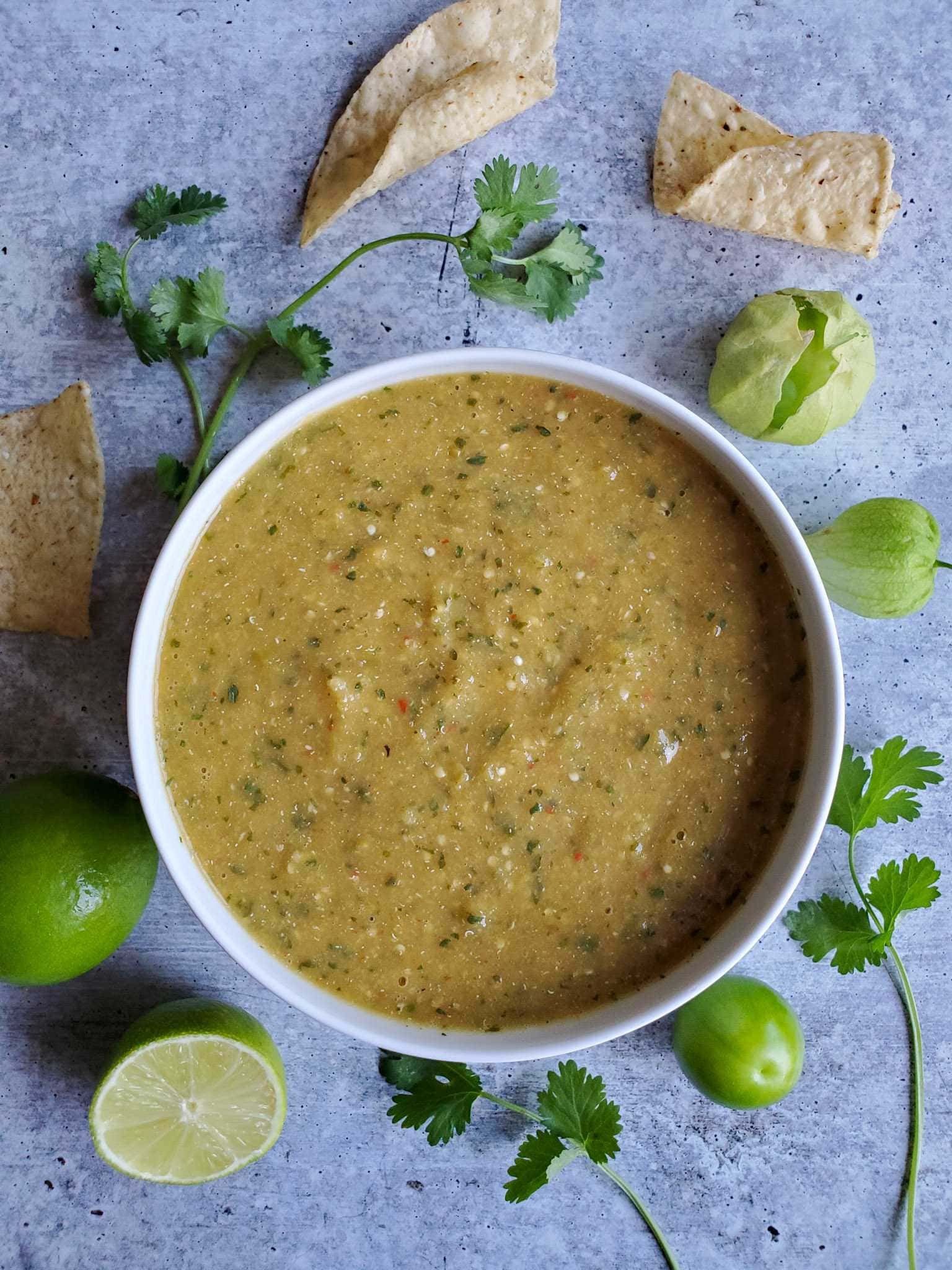 A white ceramic bowl full of roasted tomatillo salsa verde is featured. There are specks of cilantro, chili pepper, and seeds from the pepper visible. A whole lime, half a lime, fresh tomatillos, sprigs of cilantro and tortilla chips are scattered around the outside of the bowl.