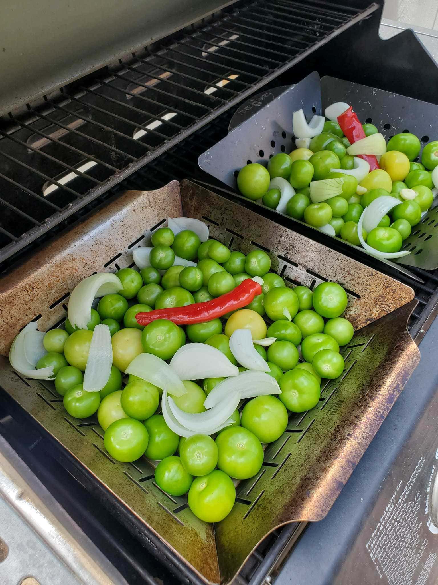 The inside of a BBQ grill is shown with two stainless steel grilling baskets sitting atop the grill. Each basket is full of fresh tomatillos, quartered onions, garlic cloves, and a red chili pepper.