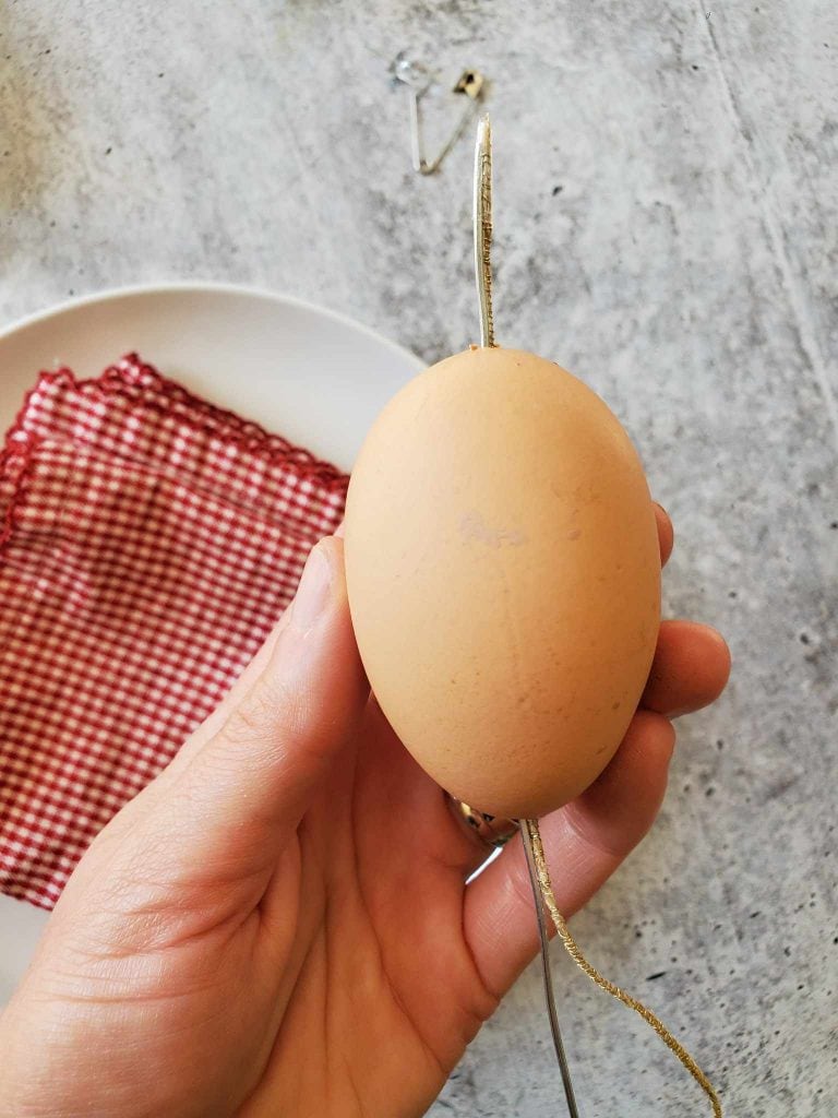 DeannaCat is holding the egg and feeding the twine and wire through the center of the egg, in and out of each hole on both ends. In the background lies a red and white striped cloth on a white plate.