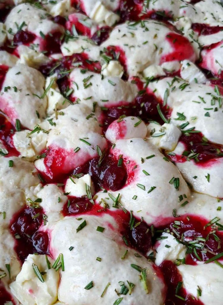 An unbaked loaf of sourdough focaccia is featured with pockets of cranberry sauce, chunks of white soft cheese, and fresh chopped rosemary set against the pillowy white dough. Into the oven it will go!