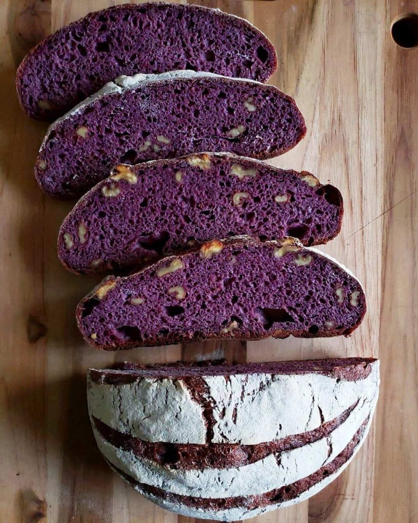 A sourdough boule loaf has been cut in half on a wood cutting board. One half of the loaf has been cut into four slices revealing a beautifully stark purple color inside with pockets of walnuts. The bread has been dyed with black nebula carrots which turned the entire loaf dark purple. The other half of the loaf has streaks of purple in the top from where the loaf was scored set against a floury white crust.