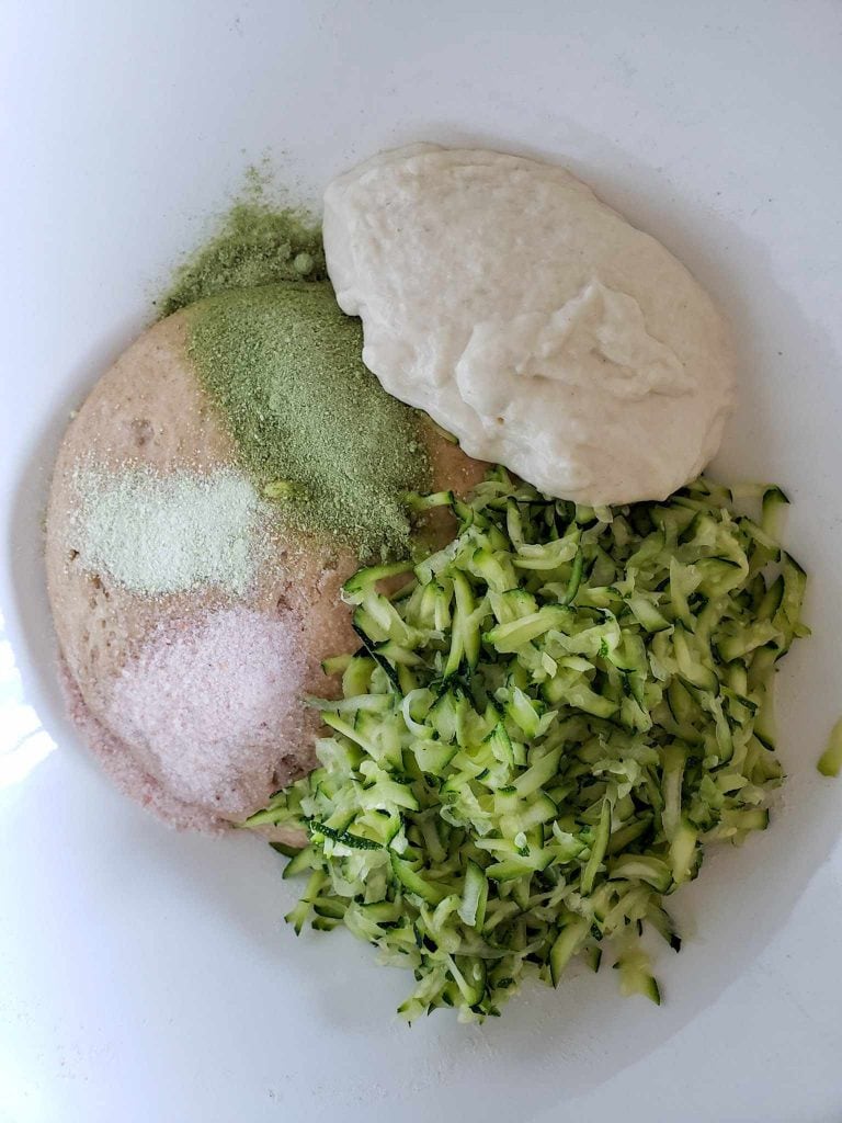 A close up of the inside of a white bowl, there is a dough ball in the very bottom with pink salt, a light greenish powder, and a darker green powder in three distinct lines across the top left half of the dough, the right half of the dough is covered equally by shredded zucchini on the bottom and sourdough starter on the upper right.