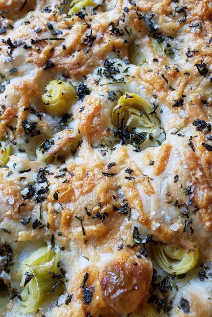 A close up of the top of a freshly baked sourdough focaccia loaf. Chopped herbs, melted cheese, and sliced artichoke hearts were the sourdough add-ins. The loaf is a mixture of golden brown crust, gooey white cheese, green artichoke hearts, and speck of chopped herbs.