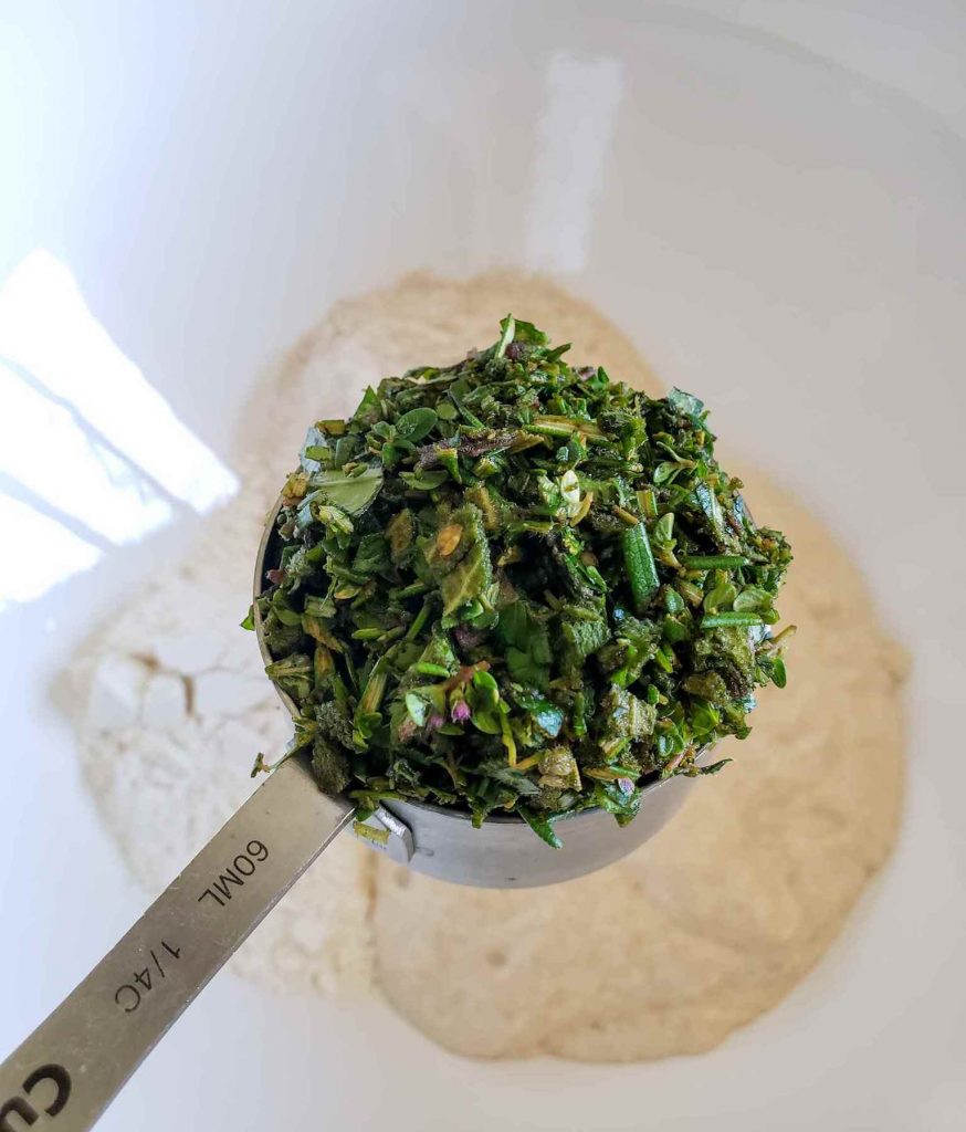 A stainless steel 1/4 cup measuring cup is positioned over a bowl of flour and sourdough starter. The measuring cup is loaded with fresh chopped herbs which are a great sourdough add-in.