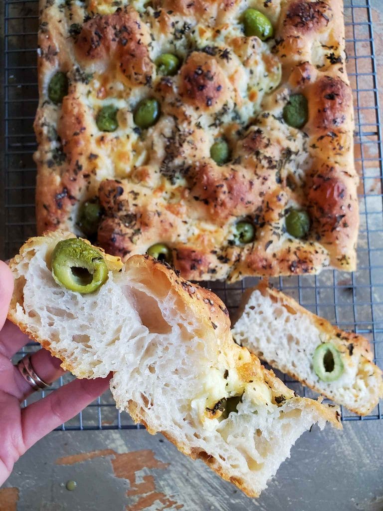 A slice of focaccia is featured, its fluffy interior is visible with air pockets here and there. A green olive is slightly blistered and shrunken from its original size due to the baking process. A pocket of cheese is on the opposite end of the slice. Below lies the remainder of the loaf of focaccia bread on a wire cooling rack. The top of the loaf is dotted with green olives, pockets of cheese, and fresh chopped herbs. Its bubbly surface is golden brown.