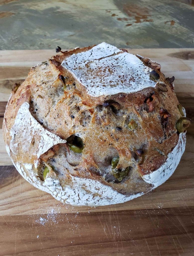 A sourdough boule loaf is sitting atop a wooden cutting board. The crust has sections of white flour adhered to the outside while the remainder of the loaf is a dark golden brown. Green olives and walnuts are bursting out from the inside of the loaf as they were the sourdough add-ins of choice.