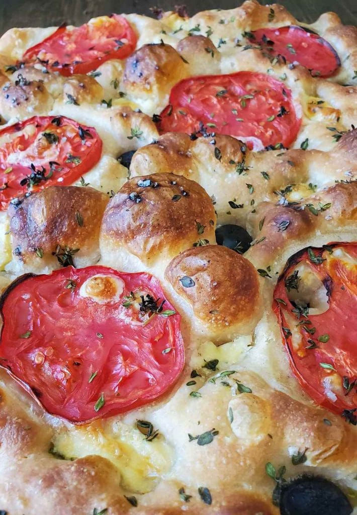 A close up of baked sourdough focaccia, with a light brown bubbly surface, flecks of little green thyme leaves, slices of round tomatoes, and black olives pressed into the dimples on top of the dough.