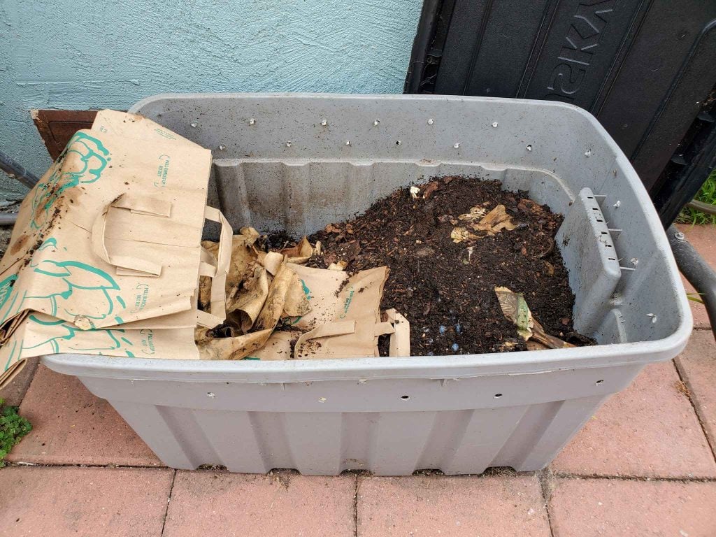 A grey tote being used for a worm bin. Holes are drilled into the tote around the top 3 inches of the tote for airflow. The tote contains a mixture of material that makes up the worm bedding, castings, and food. Some brown paper bags are off to the side of the tote which are used to cover the top of the bedding material when not in use.