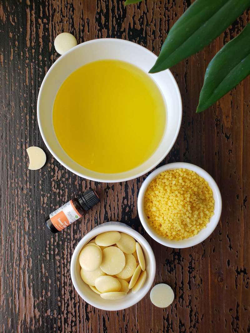 A birds eye view of three white bowls of varying size. The largest bowl contains some type of oil, another smaller bowl contains beeswax pastilles while the final bowl contains cocoa butter wafers. A small bottle of essential oil and a couple cocoa butter wafers garnish the three bowls.