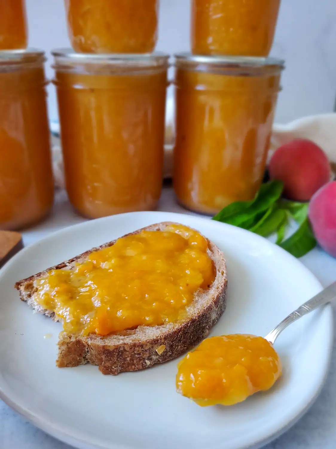A white ceramic plate has a slice of toast that is covered in preserves. A silver spoon sits next to it full of the jam. Full jars of jam are just beyond along with a few peaches and even some leaves from the tree.