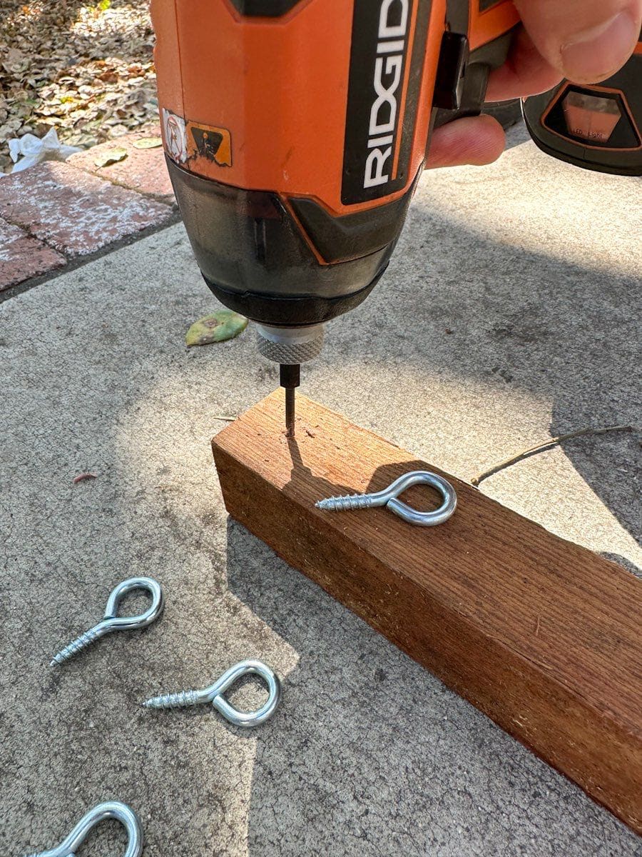 A drill is being used to add pilot holes to the ends of the 2x2 pieces of wood, 4 eye hooks are laying around the piece of wood. The eye hook will be screwed into each one of the holes.