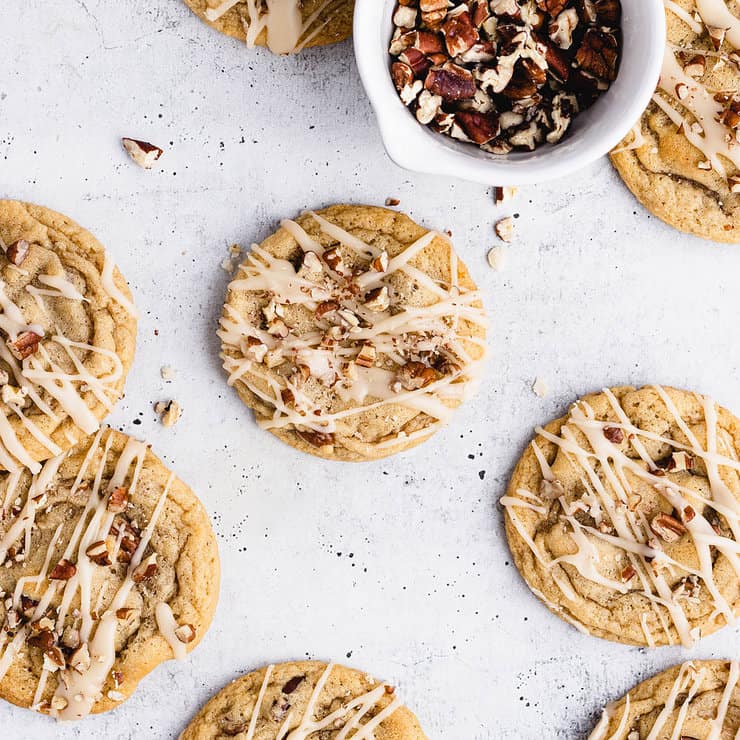 Three maple cookies on a small parchment paper-lined silver cooling rack. Around them are a glass of milk, a bowl of glaze, plates and a small bowl of pecans.